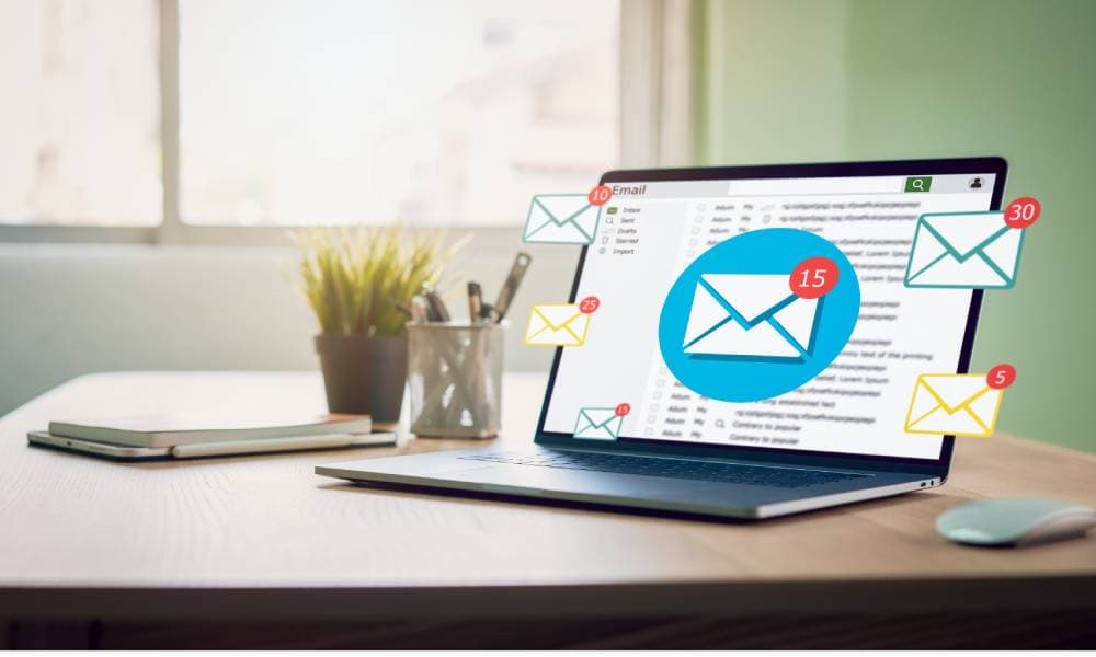 A laptop sitting on a desk with an email inbox on the screen. Colored email alert icons are scattered across the screen.