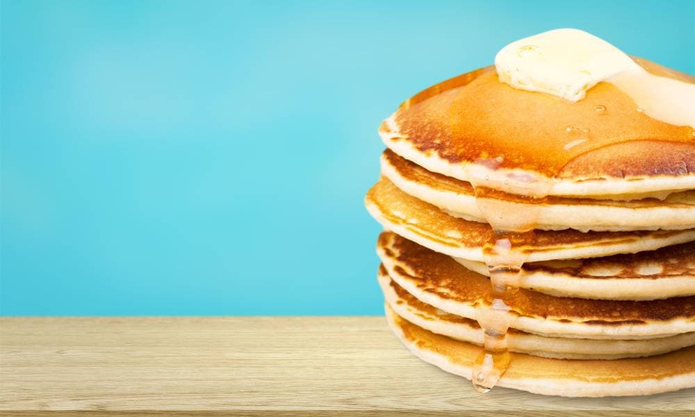 A stack of fluffy pancakes. Syrup is melting down the pancakes and the first pancake in the stack has a pat of butter.