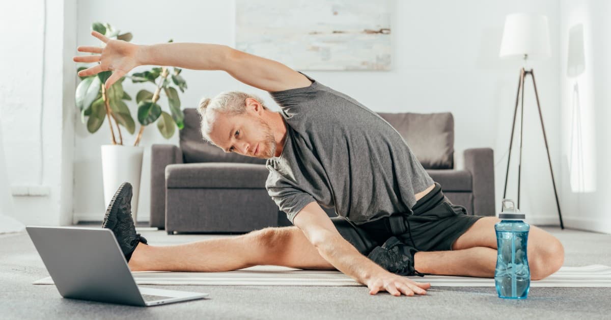 A man is sitting on a yoga mat on the floor and stretching. He is wearing shorts and a t-shirt and looking at his laptop.