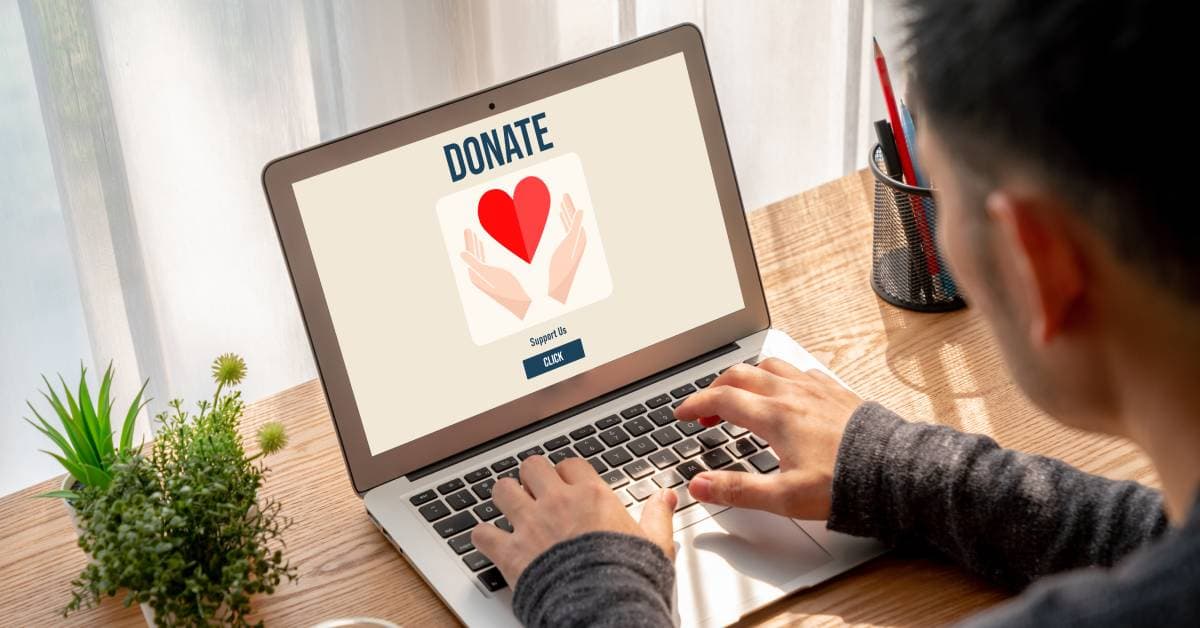 A man sitting at a desk and typing on his laptop. The screen is displaying a donation page with two hands around a red heart.