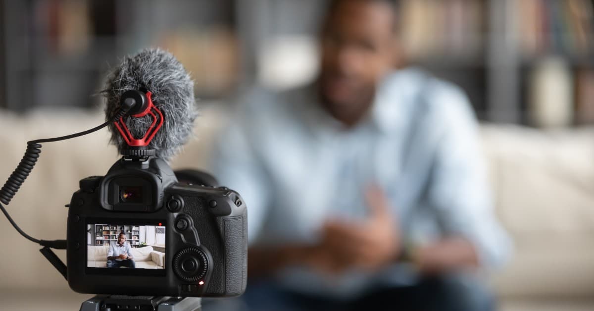 A close-up view of a black video camera focusing on a man sitting on a couch. The man is blurred in the background.