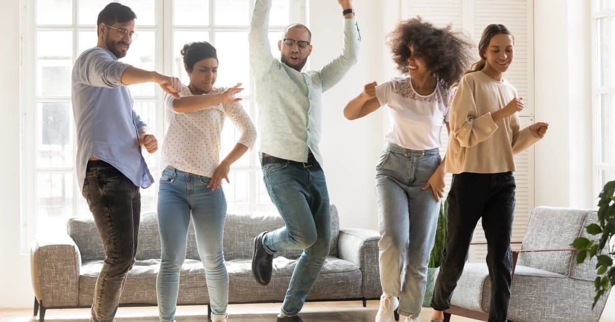 A group of five people are smiling and dancing in a bright indoor room with a gray couch and armchair.