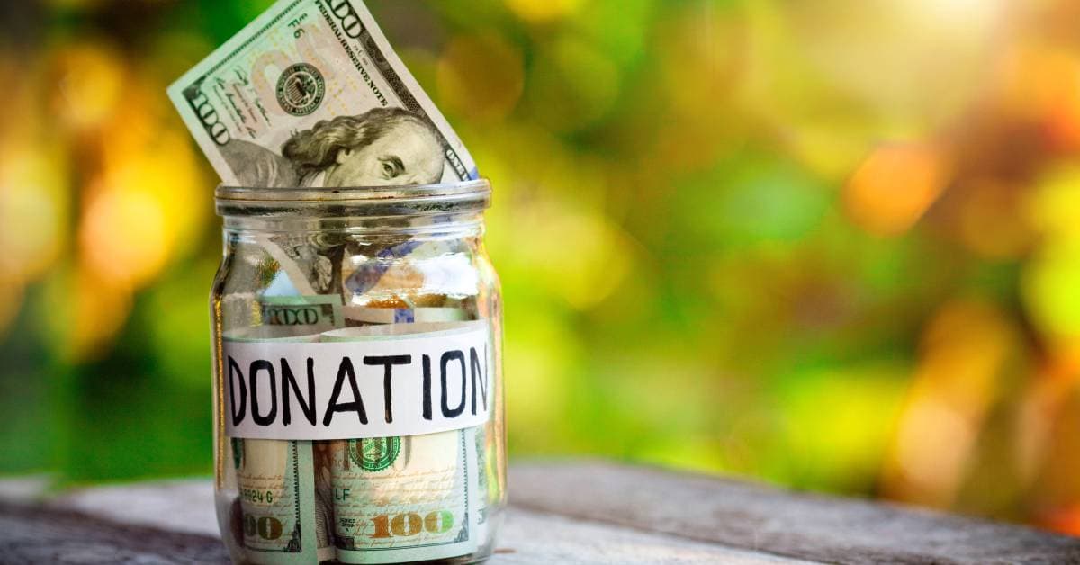A glass jar filled with several one hundred dollar bills. The jar has a label that says "donation" in black text.