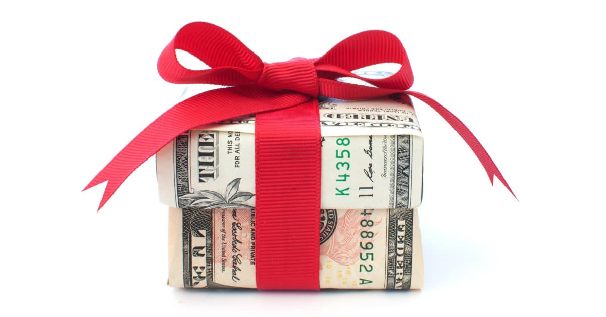 A small box is wrapped in United States paper currency and tied with a red fabric bow. The box is on a white background.