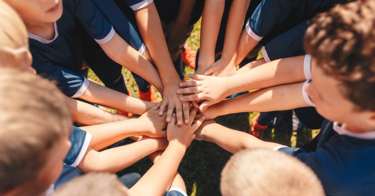 A group of kids wearing blue and white uniforms are standing outside in a circle and stacking their hands.