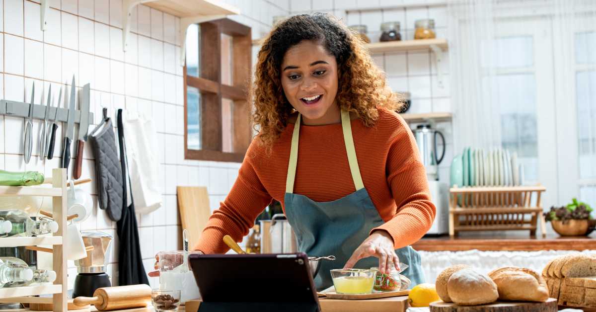 A young woman is wearing an apron and cooking in her kitchen while looking at a tablet in a black case.