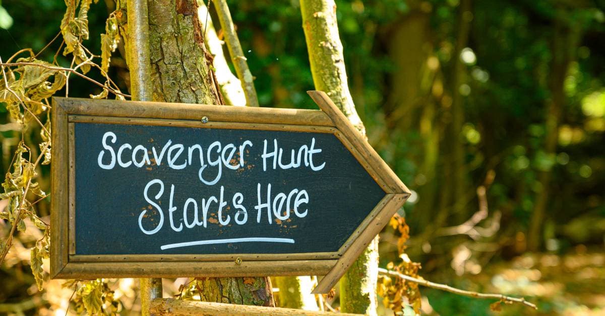 A wood sign in the shape of an arrow says "Scavenger Hunt Starts Here" in white text. The sign is affixed to a tree.