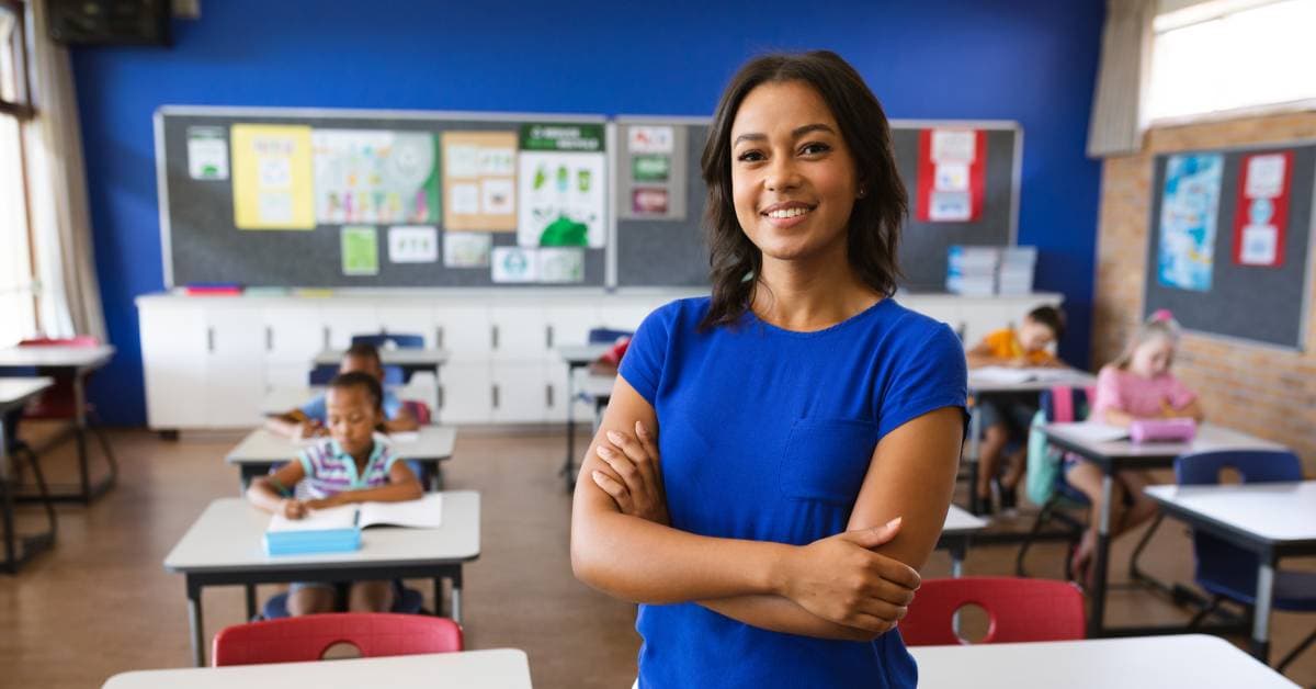 A woman wearing a blue shirt and white pants is standing in a classroom. Students are sitting at desks behind her.